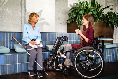Eine Frau mit Blindestock und eine Frau im Rollstuhl sitzen nebeneinander und unterhalten sich