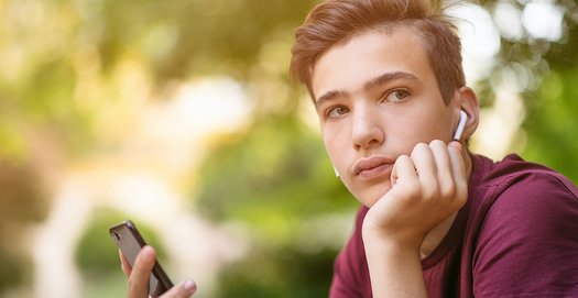 Ein Jugendlicher mit Kopfhörern im Ohr und Handy in der Hand schaut nachdenklich